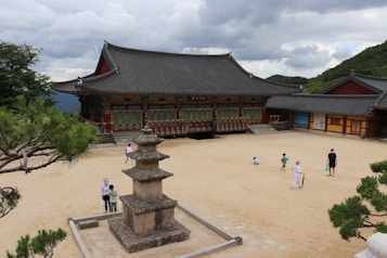 A traditional Asian temple with ornate architectural elements, including a tiled roof and colorful woodwork, surrounded by a spacious courtyard. Visitors, including adults and children, are exploring the area. A stone pagoda is prominently located in the foreground, bordered by neatly trimmed pine trees. The setting is serene, with hills and a cloudy sky in the background.