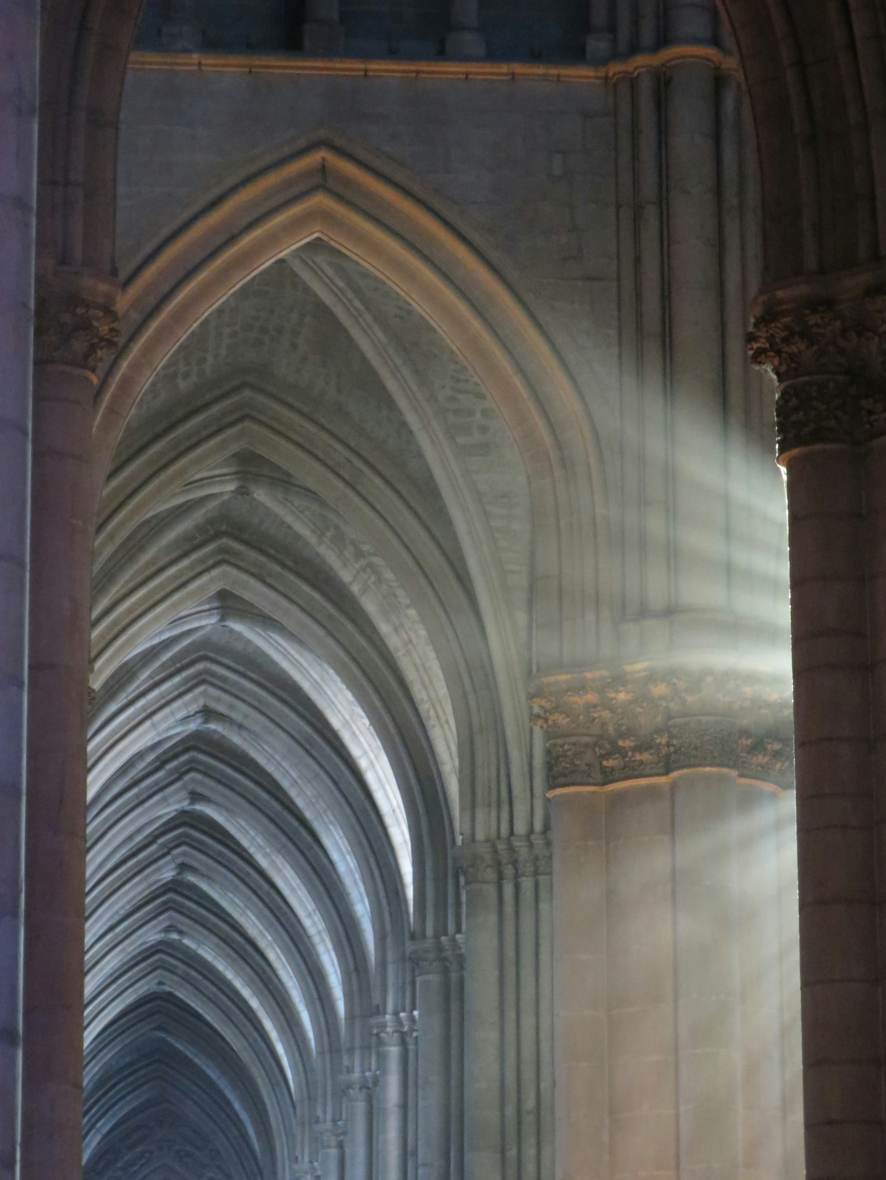 Spiritual rays of sunlight shine in a window of the Reims Cathedral. The light illuminates the first in a long row of gothic arches in the interior of the church.
