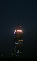 A tall, illuminated skyscraper stands against a dark night sky. A digital clock displaying the time 6:32:01 is prominently lit at the top. The building is structured with multiple floors, each with lights, and the word 'LATINOAMERICANA' is visible below the clock.