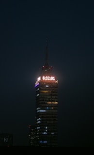 A tall, illuminated skyscraper stands against a dark night sky. A digital clock displaying the time 6:32:01 is prominently lit at the top. The building is structured with multiple floors, each with lights, and the word 'LATINOAMERICANA' is visible below the clock.