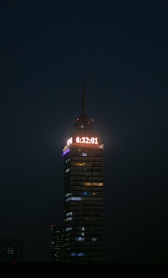 A tall, illuminated skyscraper stands against a dark night sky. A digital clock displaying the time 6:32:01 is prominently lit at the top. The building is structured with multiple floors, each with lights, and the word 'LATINOAMERICANA' is visible below the clock.