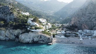A picturesque coastal scene featuring white buildings nestled on a rocky hillside covered in lush greenery. The foreground shows a clear, blue-green sea lapping against the shore of a pebbled beach with empty sunbeds. Towering cliffs frame the background, rising majestically to meet a misty sky.