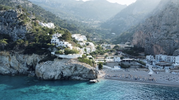 A picturesque coastal scene featuring white buildings nestled on a rocky hillside covered in lush greenery. The foreground shows a clear, blue-green sea lapping against the shore of a pebbled beach with empty sunbeds. Towering cliffs frame the background, rising majestically to meet a misty sky.
