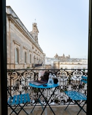 A charming view from a balcony overlooking a historic European town. The balcony features ornate, black wrought iron railings with a round blue table and two matching blue chairs. On the table are a laptop, a water bottle, and a mug. In the background, there is a large stone building with arched windows and intricate architectural details, complemented by other historic buildings.