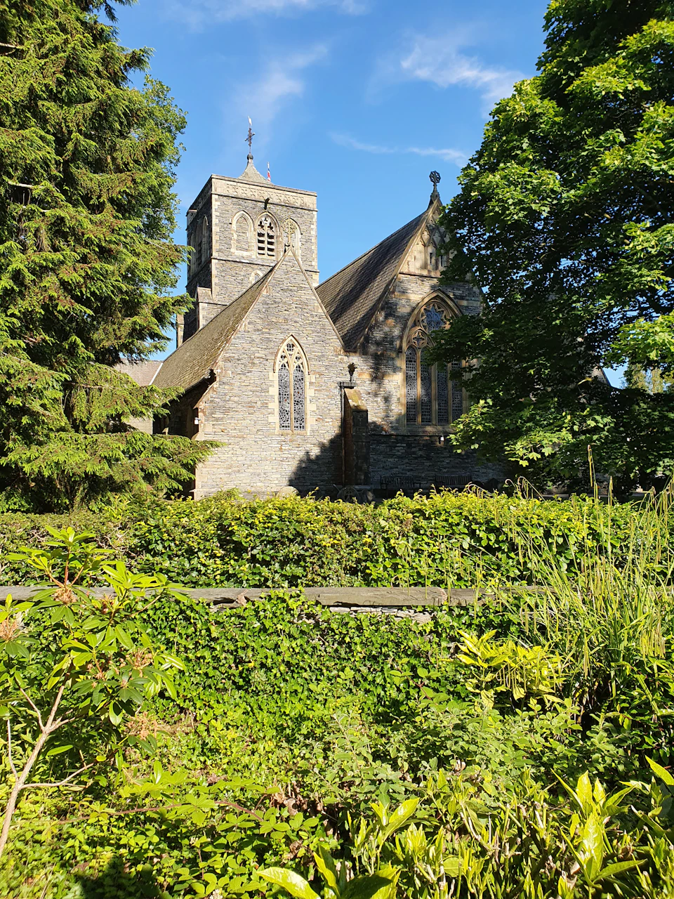 The image depicts a serene scene featuring a stone church with a tall tower and a steeple, nestled amidst lush greenery. The church is surrounded by an abundance of trees and bushes, creating a sense of tranquility and peace. The perspective of the photo is from ground level, looking up at the church, which enhances its grandeur and prominence in the landscape.
