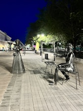 Metal sculptures of a woman and a seated artist are placed on a paved street under evening lighting. The woman appears to be posing with a flowing dress, while the artist holds a frame, as if capturing the scene. Trees and streetlights line the pathway, with a building visible on the left and people walking in the distance. The scene has a calm, artistic vibe.