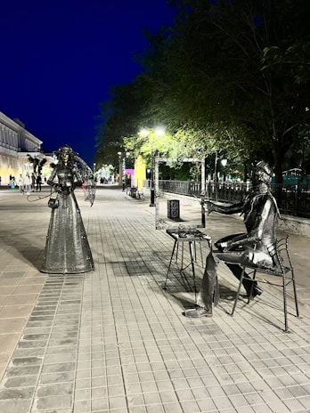 Metal sculptures of a woman and a seated artist are placed on a paved street under evening lighting. The woman appears to be posing with a flowing dress, while the artist holds a frame, as if capturing the scene. Trees and streetlights line the pathway, with a building visible on the left and people walking in the distance. The scene has a calm, artistic vibe.