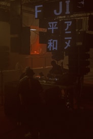 A dimly lit room features several electronic screens displaying text in Roman and Japanese characters. The area appears to be a DJ booth or music venue with equipment like turntables and speakers. There are silhouettes of people interacting or observing the performance, creating a shadowy ambiance.