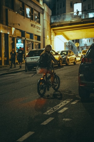 A cyclist wearing a helmet rides along a dimly lit urban street at night. The scene includes parked cars lining the roadway and a modern bridge or overpass illuminated by artificial light. Pedestrians walk along the sidewalk, and buildings with illuminated windows are visible in the background.