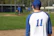 A smiling young baseball player in a navy jersey throwing a ball on a sunny field.