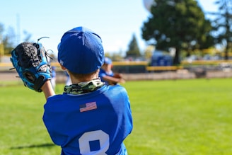 A child wearing a blue baseball uniform with the number 8 and a small American flag on the back is on a baseball field. The child is holding a blue baseball glove and appears to be catching a ball. Another player, slightly out of focus, is seen in the background. The field is green with trees and a fence surrounding it.