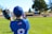 A child wearing a blue baseball uniform with the number 8 and a small American flag on the back is on a baseball field. The child is holding a blue baseball glove and appears to be catching a ball. Another player, slightly out of focus, is seen in the background. The field is green with trees and a fence surrounding it.