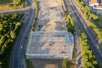 Aerial view of a large construction site with a metal framework of a building under construction. Surrounding the site are several roads with vehicles and dense greenery. The sun casts long shadows, indicating it might be early morning or late afternoon.