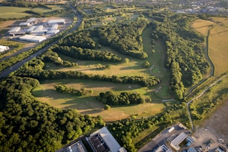 An aerial view of a golf course surrounded by dense, lush greenery. The course features several fairways, sand traps, and a variety of trees creating natural barriers. Adjacent to the golf course are industrial buildings and a main road, juxtaposing the natural environment against human development.