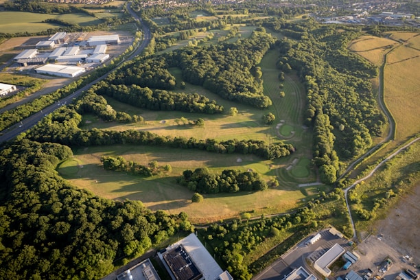 An aerial view of a golf course surrounded by dense, lush greenery. The course features several fairways, sand traps, and a variety of trees creating natural barriers. Adjacent to the golf course are industrial buildings and a main road, juxtaposing the natural environment against human development.
