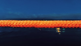 A vibrant orange rope is prominently displayed against a dark blue, possibly night sky or ocean backdrop. The texture of the rope is detailed, with visible fibers suggesting a strong weave. Below, there are faint reflections or lights against a dark background, adding depth and a sense of mystery.