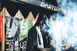 A man wearing a cap is silhouetted against a hazy, smoke-filled background. He is surrounded by various posters with bold text, promoting different products. The scene seems to take place in an urban setting, possibly at night, given the lighting and atmosphere.