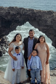 A family of five stands together posing for a photo with a rocky coastal background. The family consists of two adults and three children dressed in formal attire, standing on a cliff with waves crashing against the rocks behind them.