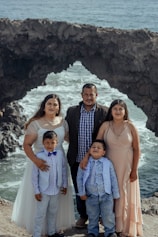 A family of five stands together posing for a photo with a rocky coastal background. The family consists of two adults and three children dressed in formal attire, standing on a cliff with waves crashing against the rocks behind them.