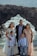 A family of five stands together posing for a photo with a rocky coastal background. The family consists of two adults and three children dressed in formal attire, standing on a cliff with waves crashing against the rocks behind them.