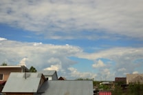 A landscape featuring several houses with metal roofs under a partly cloudy sky. The sky is vast and blue, dotted with fluffy white clouds. The houses are surrounded by greenery, and there is a bright, peaceful ambiance.