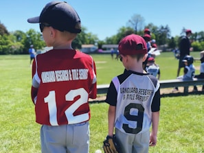 Two young children in baseball uniforms are standing on a grassy baseball field. The child on the left is wearing a red jersey with the number 12, while the child on the right is wearing a white and black jersey with the number 9. Both have baseball caps on, and the child on the right is holding a baseball glove. Other children and some adults can be seen in the background, participating in what appears to be a baseball game.