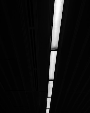 A long, linear fluorescent light fixture is embedded in a dark ceiling, casting a bright, white light that contrasts sharply against the surrounding darkness. The ceiling features a repetitive pattern with lines that run parallel to the light fixture.