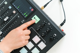 A hand pressing the record button on a digital audio mixer, which includes sliders, knobs, and a display screen showing sound levels.