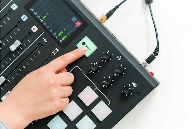 A hand pressing the record button on a digital audio mixer, which includes sliders, knobs, and a display screen showing sound levels.