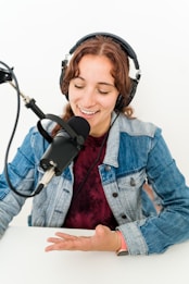 A person wearing headphones is speaking into a professional microphone. They are sitting at a white table and appear to be engaged in podcast recording or broadcasting. The person is wearing a denim jacket over a dark top.