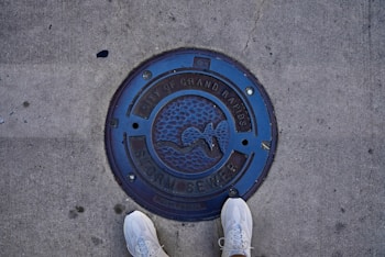 A circular manhole cover labeled 'City of Grand Rapids Storm Sewer' is embedded in a concrete sidewalk. The cover is dark blue with a textured design featuring a bird in flight. Two feet wearing white sneakers are visible at the bottom of the image, standing on the pavement.