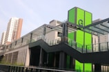 A modern urban structure with a prominent green panel featuring the letter 'G'. The building includes sleek glass and metal architectural elements. In the background, a multi-story residential building is visible under a clear sky.