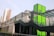 A modern urban structure with a prominent green panel featuring the letter 'G'. The building includes sleek glass and metal architectural elements. In the background, a multi-story residential building is visible under a clear sky.