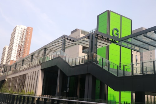 A modern urban structure with a prominent green panel featuring the letter 'G'. The building includes sleek glass and metal architectural elements. In the background, a multi-story residential building is visible under a clear sky.