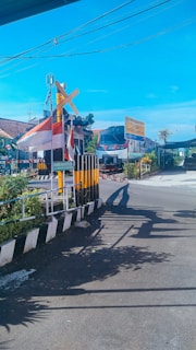A railway crossing with a train approaching in the background. The crossing is equipped with barriers and pedestrian fences, painted in black, white, and yellow stripes. There is an Indonesian flag on top of the crossing gate. The area is surrounded by greenery with houses visible on the left. Overhead, there are some power lines against a clear blue sky.