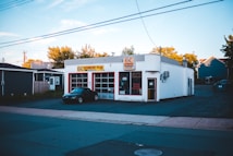 A small automotive repair shop with a distinctive red and white facade, located on a quiet street. The building is one story with large glass windows and a sign displaying the name of the business. A black car is parked in front of one of the garage doors. Tree-lined streets with houses are visible in the background, bathed in the soft light of late afternoon.