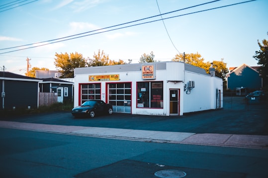A small automotive repair shop with a distinctive red and white facade, located on a quiet street. The building is one story with large glass windows and a sign displaying the name of the business. A black car is parked in front of one of the garage doors. Tree-lined streets with houses are visible in the background, bathed in the soft light of late afternoon.