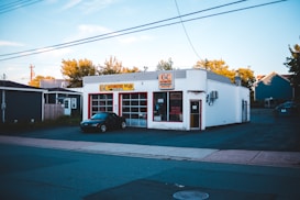 A small automotive repair shop with a distinctive red and white facade, located on a quiet street. The building is one story with large glass windows and a sign displaying the name of the business. A black car is parked in front of one of the garage doors. Tree-lined streets with houses are visible in the background, bathed in the soft light of late afternoon.