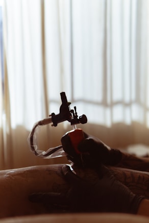 A tattoo artist, wearing black gloves, is applying a tattoo with a tattoo machine on a person's skin. The room is softly lit, and draped curtains in the background create a serene and focused atmosphere.