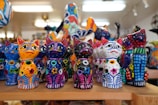 A collection of vividly colored, handcrafted cat figurines arranged on a wooden shelf. Each figure features bright patterns and designs that include stars and dots in multiple colors. The background is softly blurred, suggesting a shop or display setting.