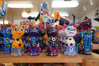 A collection of vividly colored, handcrafted cat figurines arranged on a wooden shelf. Each figure features bright patterns and designs that include stars and dots in multiple colors. The background is softly blurred, suggesting a shop or display setting.