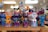 A collection of vividly colored, handcrafted cat figurines arranged on a wooden shelf. Each figure features bright patterns and designs that include stars and dots in multiple colors. The background is softly blurred, suggesting a shop or display setting.