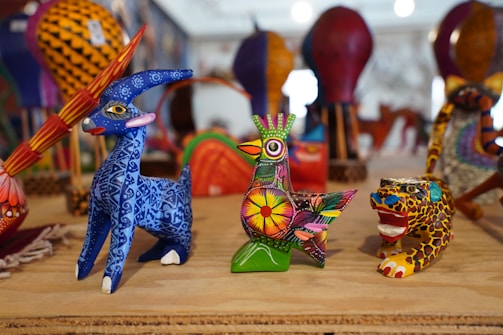 Intricately painted wooden animal figurines, including a blue deer, a colorful bird, and a jaguar, arranged on a wooden surface with various other vibrant crafts in the background.