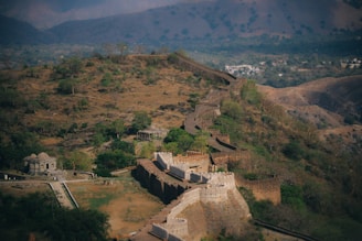 A sprawling, historic fortress stretches across rugged terrain, winding through a lush landscape dotted with trees. The stone walls, featuring large bastions and crenellations, are well-preserved, offering a glimpse into past architectural ingenuity. The background showcases rolling hills and a distant settlement, emphasizing the fortress's strategic location.