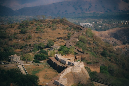 A sprawling, historic fortress stretches across rugged terrain, winding through a lush landscape dotted with trees. The stone walls, featuring large bastions and crenellations, are well-preserved, offering a glimpse into past architectural ingenuity. The background showcases rolling hills and a distant settlement, emphasizing the fortress's strategic location.
