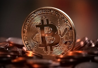 A large, golden Bitcoin coin stands prominently among a pile of similar coins, symbolizing digital currency. The background is dark, creating a strong contrast with the shiny surface of the coin.