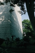 A large, textured concrete building stands adjacent to a green, leafy environment. Shadows from trees fall across the surface of the building, creating a pattern of light and dark. The trees and bushes in the foreground contribute to a natural setting, with a clear blue sky visible in the background.