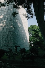 A large, textured concrete building stands adjacent to a green, leafy environment. Shadows from trees fall across the surface of the building, creating a pattern of light and dark. The trees and bushes in the foreground contribute to a natural setting, with a clear blue sky visible in the background.