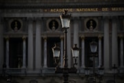 Several ornate street lamps stand in front of a classical building with tall columns and decorative sculptures. The architecture indicates a historic style with a focus on grandeur and symmetry. The building has the phrase 'Academie Nationale de Musique' inscribed at the top.