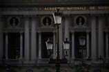 Several ornate street lamps stand in front of a classical building with tall columns and decorative sculptures. The architecture indicates a historic style with a focus on grandeur and symmetry. The building has the phrase 'Academie Nationale de Musique' inscribed at the top.
